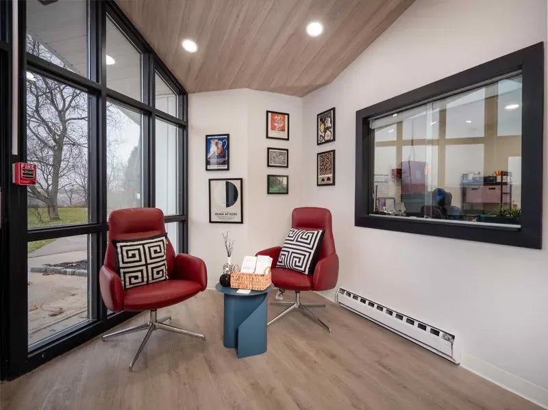 The image shows an interior space with a modern design, featuring a waiting area with a red chair, a small table, framed pictures on the wall, a window, and a door. There s a red-patterned pillow on the chair and a blue box on the table.