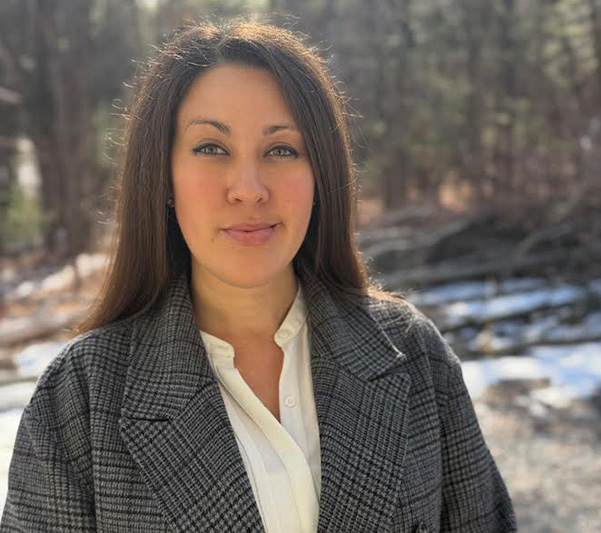 The image shows a woman standing outdoors, wearing a coat, and posing for the photo with a natural backdrop.
