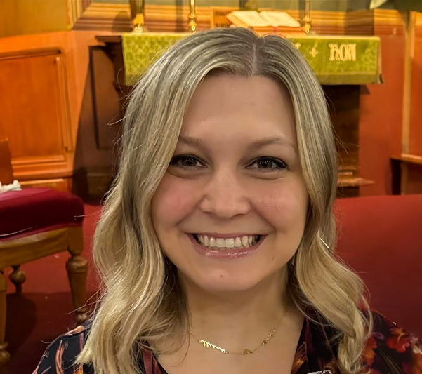 A woman with blond hair and a smiling expression, posing inside a church sanctuary with a pulpit and pews in the background.