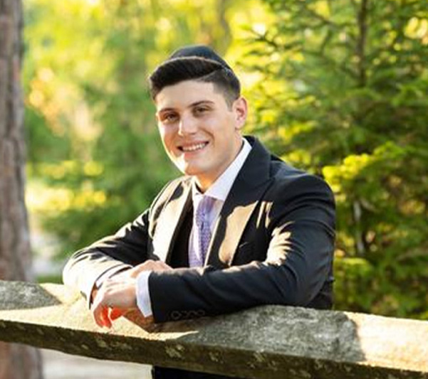 A young man wearing a suit and tie stands against a railing with a smile on his face, posing for the photograph.