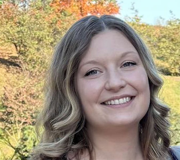The image features a woman with light hair smiling at the camera, wearing makeup, and dressed in casual attire, standing outdoors against a backdrop of autumn foliage.