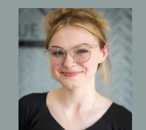 A young woman with glasses and blonde hair wearing a black top poses for a portrait.