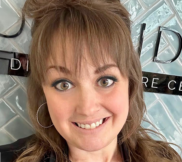 The image features a person with short hair smiling at the camera, standing against a backdrop with a partial view of a sign that reads  JUDI  and a logo resembling an eye.