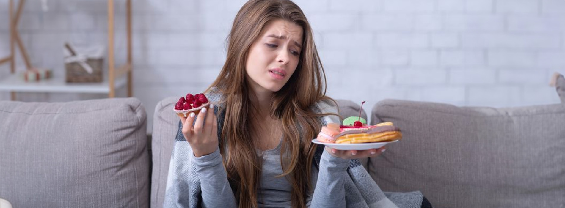 A woman sitting on a couch with her hands clasped together, wearing a gray shirt and holding a plate with two slices of cake and strawberries.