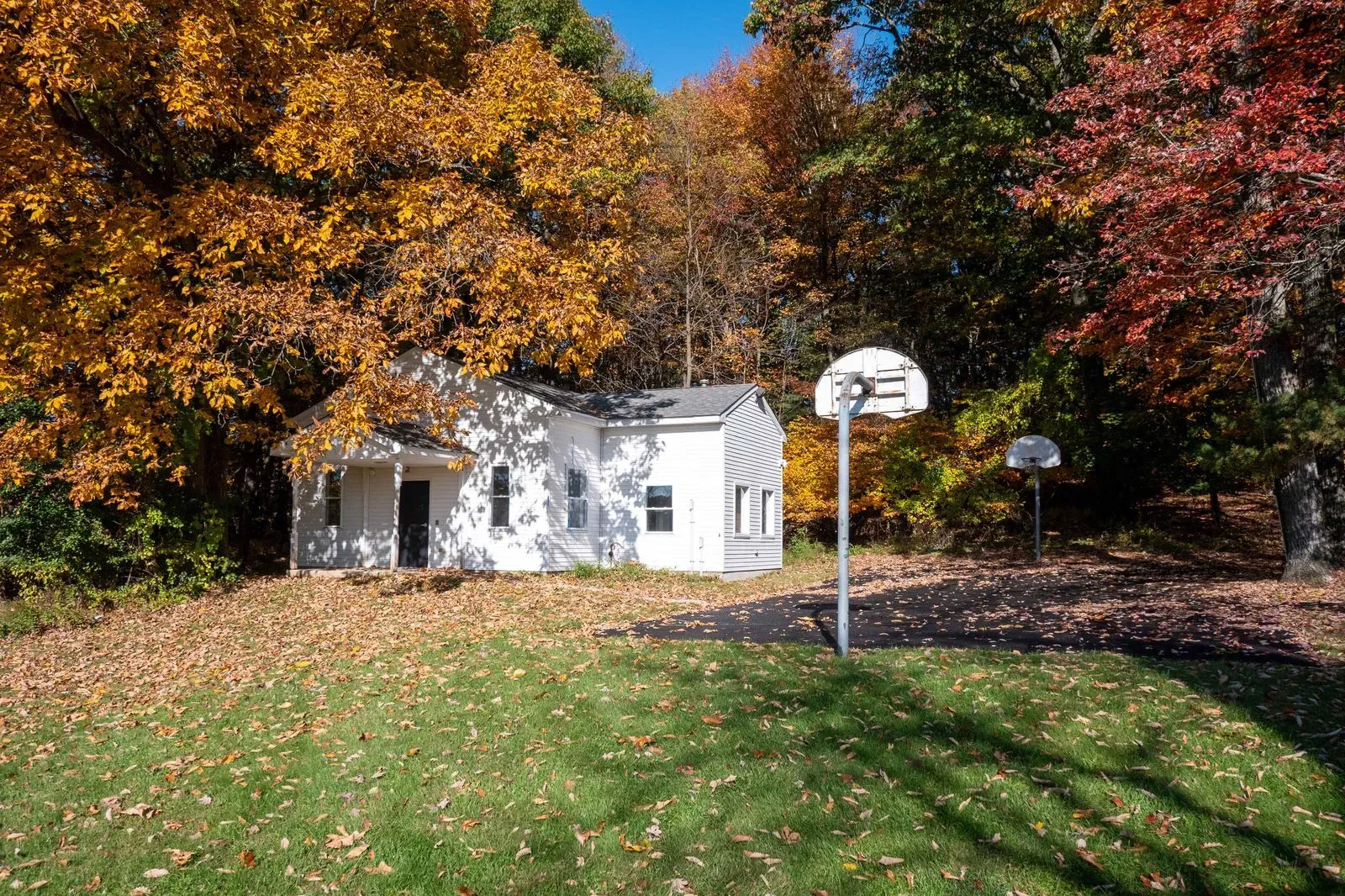 The image shows a small white house with a basketball hoop on its driveway, surrounded by autumn foliage.
